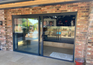 Black-framed sliding glass doors set into a rustic brick wall reflecting the outdoor swimming pool; interior features a bar with stools and framed pictures; stone patio outside; part of Glaze Ltd project