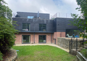 Rear view of two-storey house with red brick lower level and dark timber-clad upper level and balcony; green lawn and gabion stone wall walkway; trees partially obscuring the sides; part of Glaze Ltd project