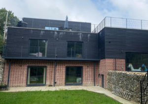 Two-level house with red brick lower level and dark timber-clad upper level with balcony; large black-framed windows; green lawn and gabion stone retaining wall; part of Glaze Ltd project