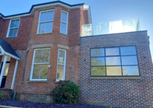 Traditional red brick house with bay window attached to modern extension featuring dark brick and a large black-framed window; reflective glass balustrade on roof terrace; part of Glaze Ltd project