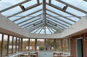 Interior of conservatory extension with glass roof supported by metal beams; windows on all sides and wooden tables and chairs inside; view of trees and garden outside; part of Glaze Ltd project